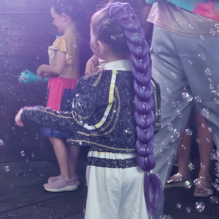 A young girl seen from behind wearing a long, thick purple braided wig, a black and white patterned jacket with gold trim, and white pants. Another child is visible in the background holding a toy bubble wand amidst floating bubbles.