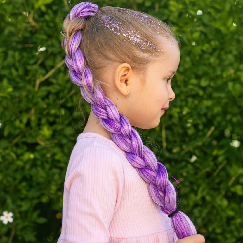 Child with purple braided hair and sparkly hair standing against a green hedge.