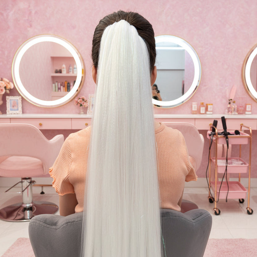 Person with long white hair sitting in a salon chair with pink walls and mirrors.