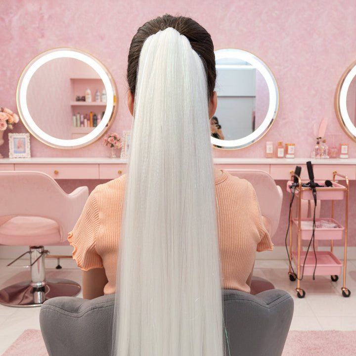 Person with long white hair sitting in a salon chair with pink walls and mirrors.