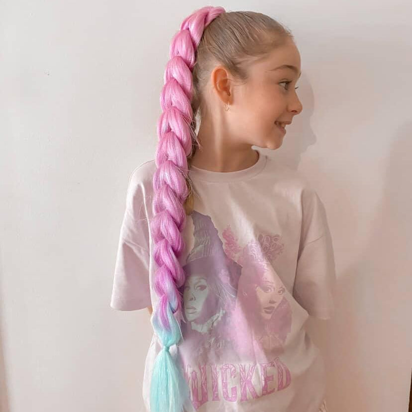 Child with pink braided dragon braid hair wearing a white shirt with graphics and checkered pants.