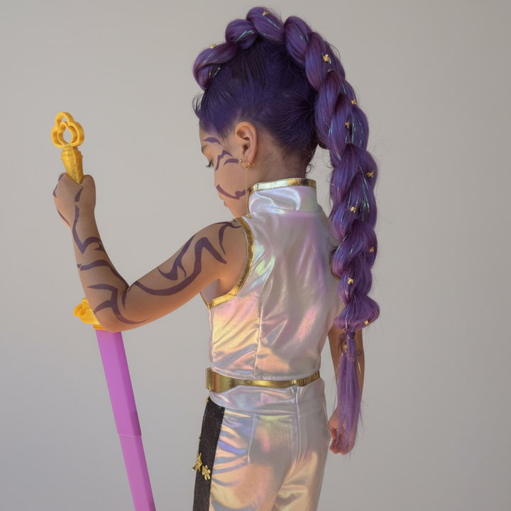 Child with purple hair in braids, body paint, and a colorful outfit holding a toy against a brick wall.