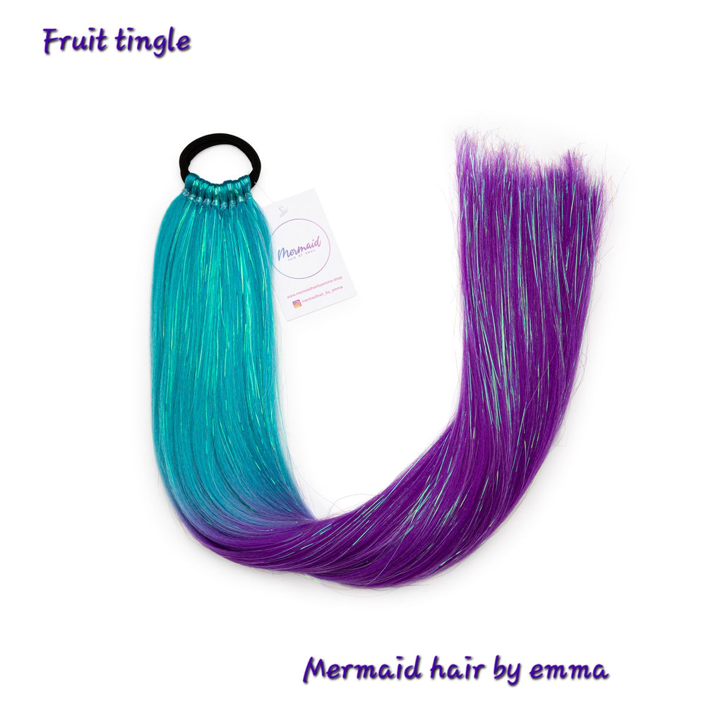 A child's mermaid-style hair extension with a gradient of turquoise to purple color, tied together with an elastic band, labeled 'Fruit tingle' by 'Mermaid hair by emma'.