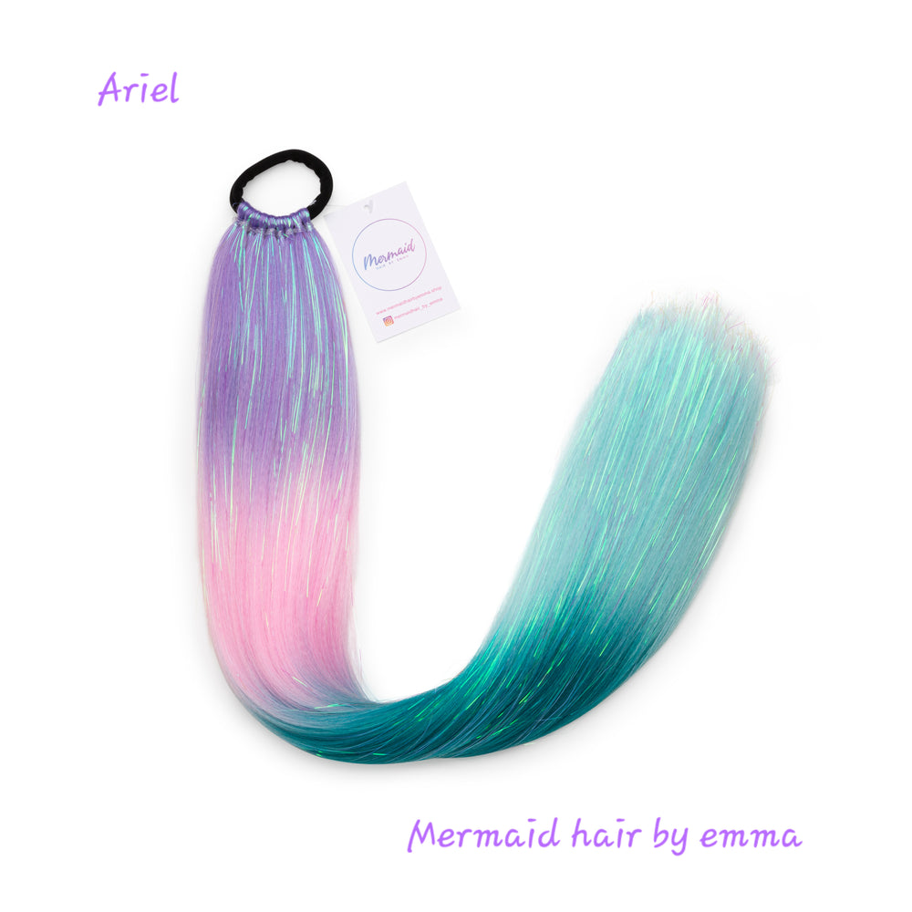 A synthetic mermaid tail hair extension with a mix of pink and blue colors, tied in a loop with an elastic band, against a white background with a label that reads 'Ariel' and 'Mermaid hair by emma'.