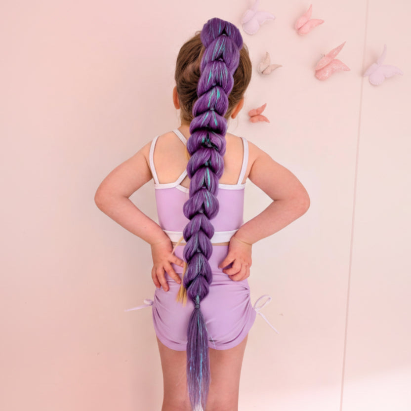 A child viewed from behind wearing a lavender romper, showcasing an exceptionally long, thick braided ponytail extension in purple with blue and yellow streaks. The background is a soft pink wall decorated with butterfly wall decals.
