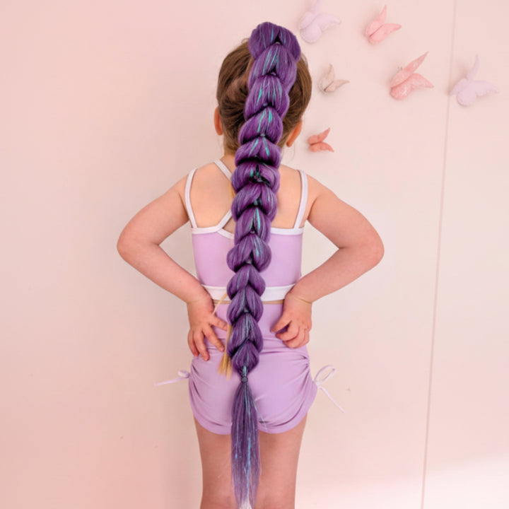A child viewed from behind wearing a lavender romper, showcasing an exceptionally long, thick braided ponytail extension in purple with blue and yellow streaks. The background is a soft pink wall decorated with butterfly wall decals.