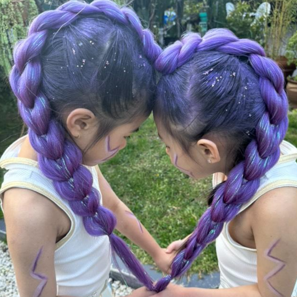 Two children outdoors with long, thick braided hair dyed in vivid purple, wearing white tank tops and looking down toward each other. The background shows grass and trees.
